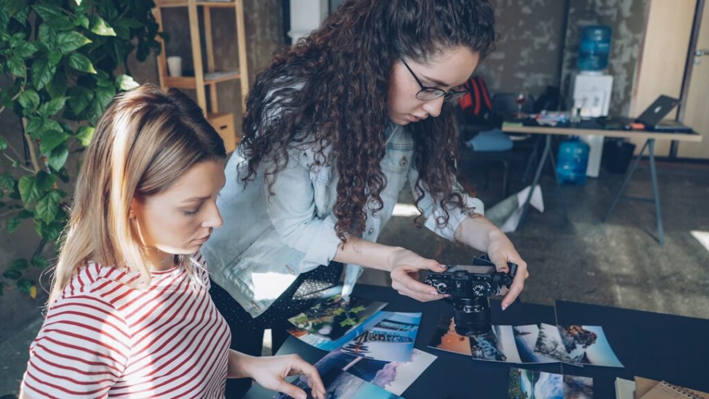 Creative colleagues placing pictures on table and shooting flat lay while talking about designing project. Top view of modern table with illustrations, notebooks and markers.