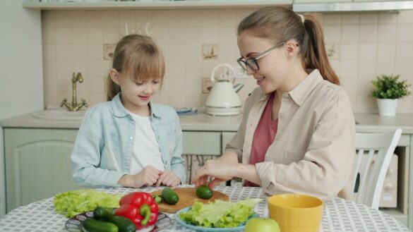 Young woman is cooking salad and feeding cute little daughter with cucumber slices in kitchen, family is talking and having good time together.