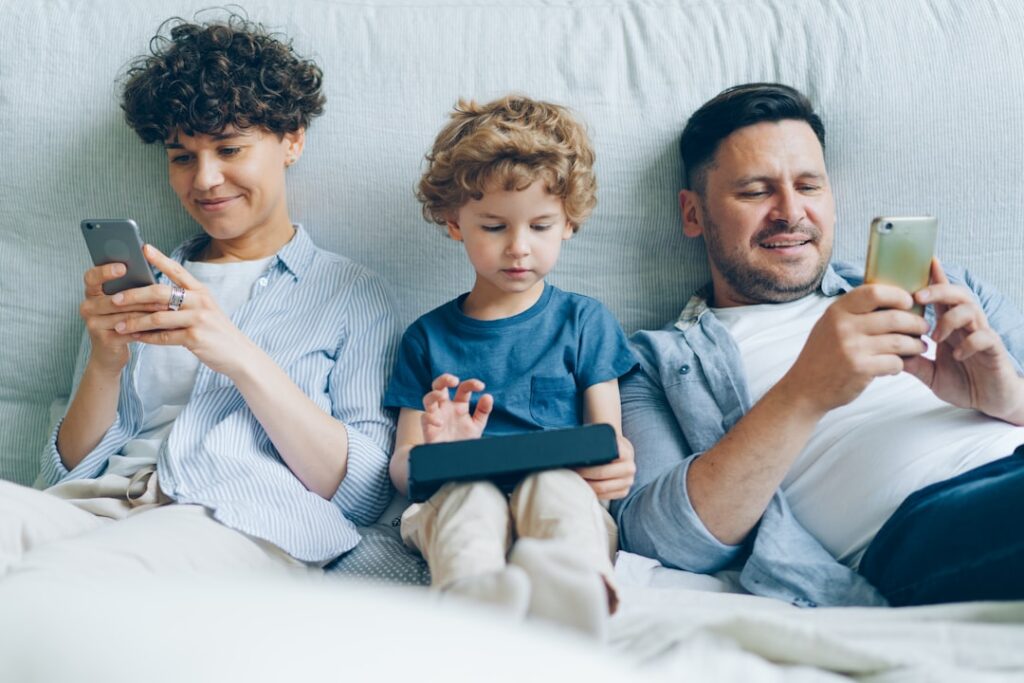 Man and woman using smartphones while kid playing game on tablet in bed at home