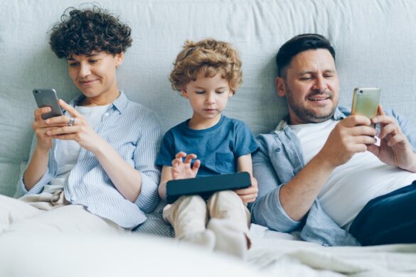 Man and woman using smartphones while kid playing game on tablet in bed at home